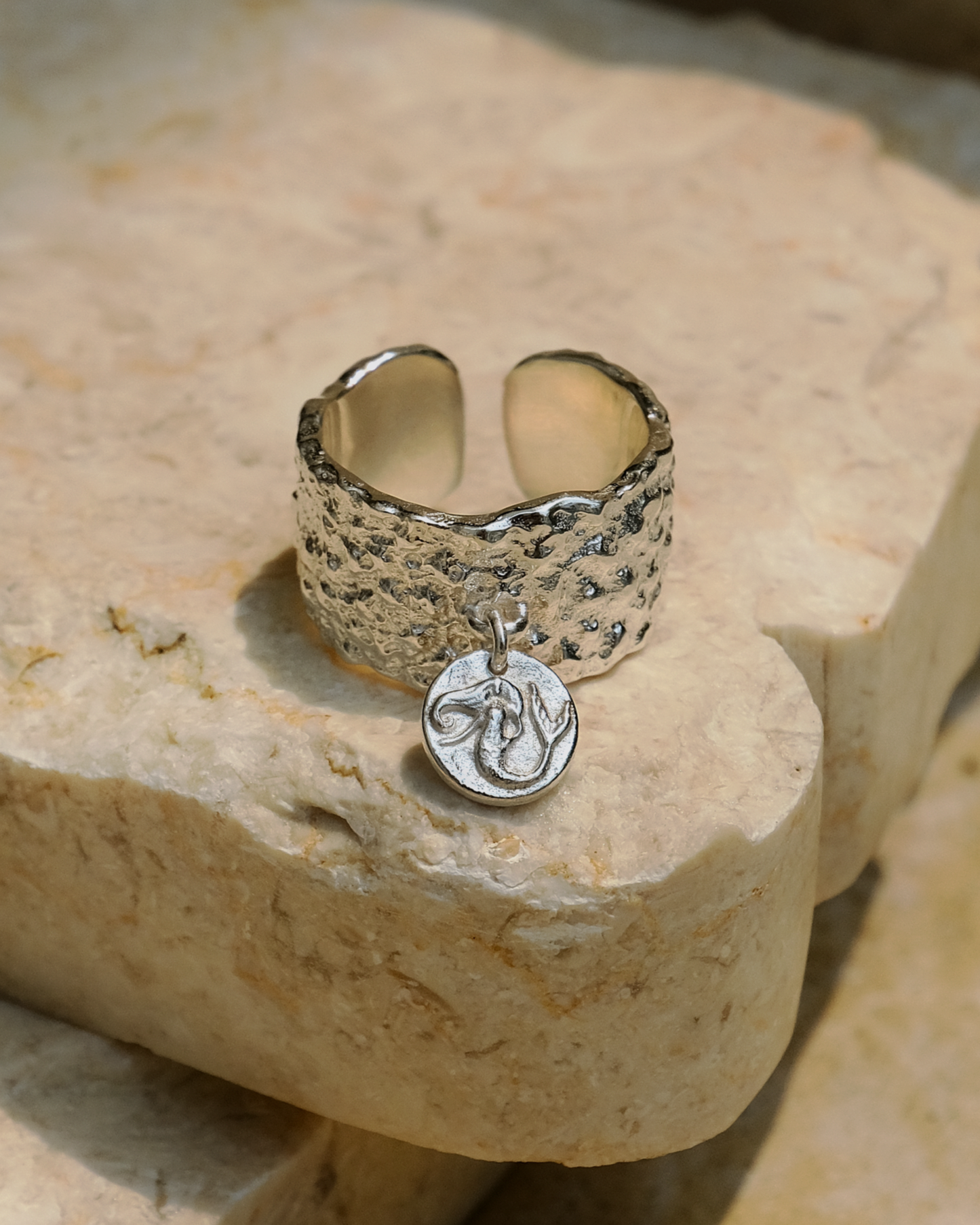 Elegant hammered silver ring with a dangling mermaid charm, featuring intricate ocean-inspired details. Handmade jewelry piece, photographed on beige stone.